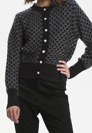 Femme portant un cardigan en maille texturée noire avec des boutons en perle et des poignets côtelés, assorti d'un pantalon noir, main sur la taille.