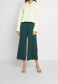 Lichtgele cropped pullover met een ritssluiting bij de kraag, gecombineerd met wijde donkergroene broeken en muntgroene sandaaltjes met bandjes.