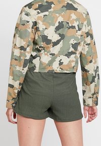 Lyhennetty kamouflage-takki beige-, vihreä- ja ruskeasävyissä, jossa napilliset hihansuut, yhdistettynä tummanvihreisiin teksturoituihin shortseihin.