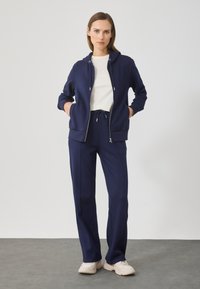 Casaco com fecho éclair azul-marinho e calças combinando, feitos de tecido suave, com cintura ajustável por cordão, acompanhado de uma t-shirt branca simples e ténis de cor clara.
