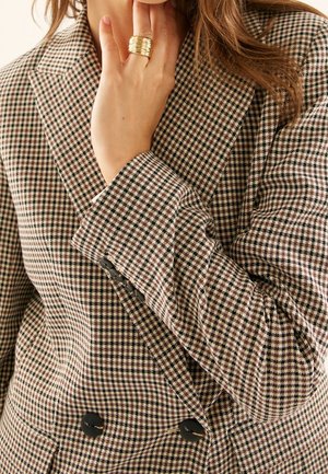 Frau trägt einen braun-schwarzen Hahnentritt-Blazer mit schwarzen Knöpfen, berührt ihr Gesicht und trägt einen goldfarbenen, strukturierten Ring am Finger.