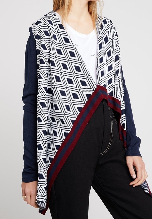 Femme portant un cardigan ouvert à motif losanges bleu marine et blanc avec des bordures bordeaux et bleu marine, sur une chemise blanche et un pantalon noir.