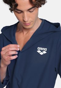 Giacca con cappuccio blu navy in morbido tessuto. Presenta una zip frontale, il logo "arena" bianco e una vestibilità rilassata che migliora la mobilità.
