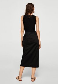 Schwarzes Kleid mit ärmellosem Design, das eine geraffte Seitenpartie und einen mittellangen Saum aufweist. Glatter Stoff und eine figurbetonte Silhouette.