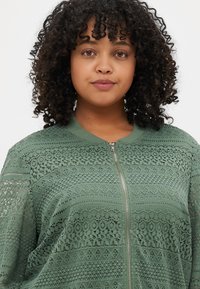 Veste bomber en dentelle vert clair avec fermeture éclair, motifs texturés, col et poignets côtelés, et coupe décontractée avec des coutures détaillées.