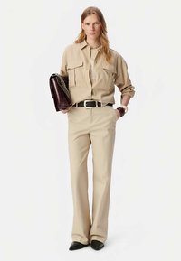 Beige overhemd met knopen en zakken, gecombineerd met bijpassende geplooide broek. Geaccentueerd met een donkere handtas en zwarte schoenen.