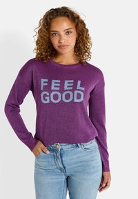 Pull à manches longues violet avec une texture pailletée portant l'inscription "FEEL GOOD" imprimée en argent. Associé à un jean bleu clair.