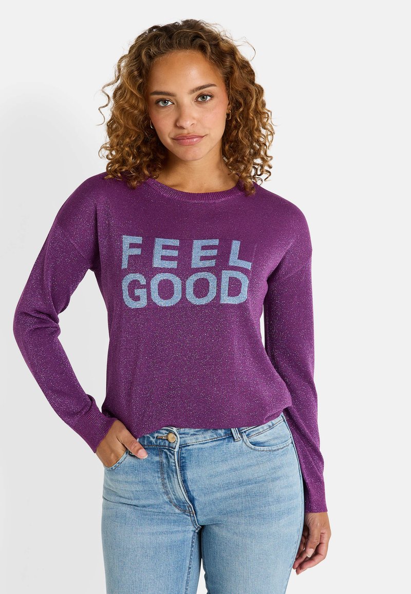 Pull à manches longues violet avec une texture pailletée portant l'inscription "FEEL GOOD" imprimée en argent. Associé à un jean bleu clair.