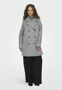 Cappotto grigio doppiopetto con grandi bottoni neri, revers ampi e un tessuto strutturato. Abbinato a pantaloni larghi neri e stivaletti alla caviglia.