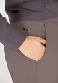 Graue Sporthose mit glatter Textur, ausgestattet mit einer Seitentasche und einem kleinen metallischen Etikett. Der Bund wirkt elastisch für zusätzlichen Komfort.