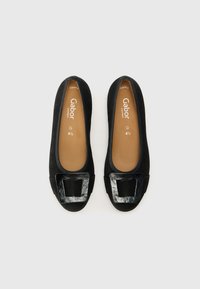 Black suede ballet flats with a glossy black square buckle detail at the toe and a tan insole with branding. Smooth finish, rounded shape.