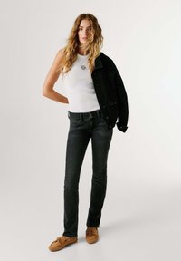 Femme portant un haut blanc sans manches, un jean noir, des mocassins beige, tenant une veste noire sur son épaule sur fond uni.