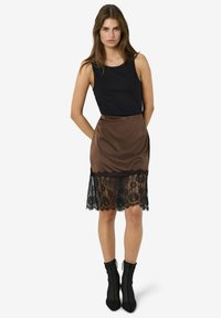 Svart ermeløs topp kombinert med et brunt satengskjørt med en svart blonderkant nederst. Modellen har på seg svarte ankelstøvler.