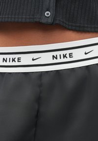 Ζωνάρι Nike σε λευκό με μαύρα γράμματα και λογότυπο, σε συνδυασμό με σκούρο γκρι ύφασμα από κάτω, με λεία, γυαλιστερή υφή.