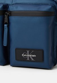 Niebieska torba z nylonu z czarnym zamkiem błyskawicznym i okuciami, z czarną etykietą z napisem "Calvin Klein" oraz wyraźnym srebrnym logo "CK".