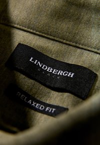 Grüner Stoff mit weicher Textur zeigt schwarze Etiketten mit weißem Text: "LINDBERGH", "BLACK" und "RELAXED FIT". Nahaufnahme.
