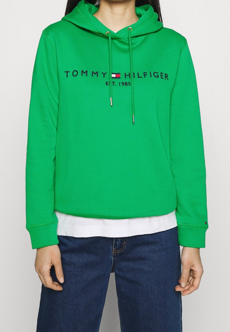 Grön hoodie i bomull med dragsnodd, med marinblå logotext och en vit, lager-på-lager skjorta under. Avslappnad passform.