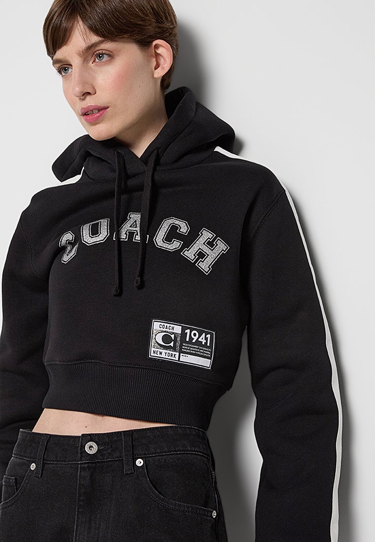 Schwarzer cropped Hoodie mit "COACH" Schriftzug in Silber, einem Etikett mit der Aufschrift "1941" und kontrastierenden Seitenstreifen. Weiches, strukturiertes Material.