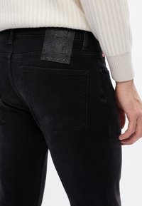 Jeans de mezclilla negra con un parche de cuero negro texturizado en la parte trasera de la cintura, con costuras visibles y un diseño elegante y ajustado.