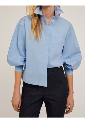 Femme portant une chemise bleu clair oversize avec des manches bouffantes et un col montant, associée à un pantalon foncé taille haute.