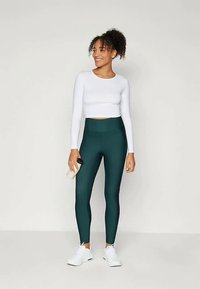 Høyt midjefestede, mørkegrønne leggings kombinert med en tettsittende hvit langermet topp. Modellen holder en vannflaske og har på seg hvite joggesko.