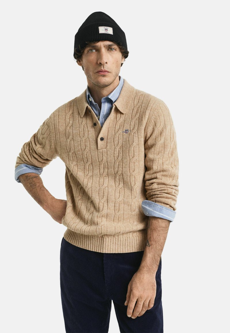 Man in een zwarte gebreide beanie, beige kabelgebreide trui over een blauwe overhemd, en donkere broek, staand met één hand op de heup.