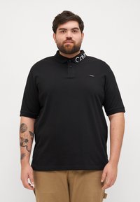 Μαύρο πόλο μπλούζα με συμπαγή σχέδιο, διαθέτει κολάρο και το λογότυπο Calvin Klein σε λευκό στην κολαρίνα. Κοντά μανίκια.