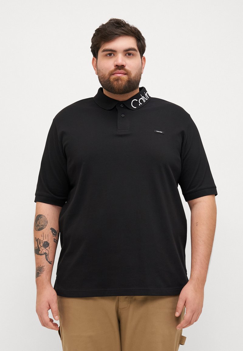 Μαύρο πόλο μπλούζα με συμπαγή σχέδιο, διαθέτει κολάρο και το λογότυπο Calvin Klein σε λευκό στην κολαρίνα. Κοντά μανίκια.