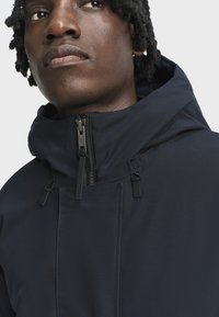 Young person with dark skin and curly hair wearing a navy blue hooded jacket with a front zipper, looking slightly upward.