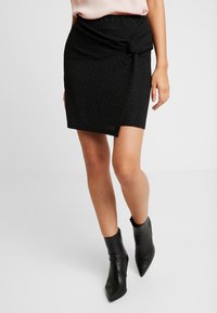 Kvinna i en texturerad svart minikjol med sidoknut och svarta spetsiga anklarstövlar, stående mot en vit bakgrund.