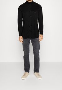 Homme portant une chemise noire à boutons manches longues, un jean gris foncé et des baskets beiges, debout devant un fond clair uni.
