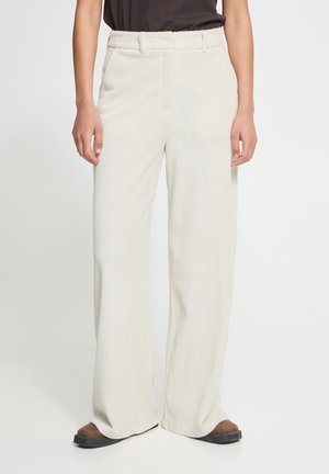 Persoon draagt off-white wijde corduroy broek met knoop- en ritssluiting, gecombineerd met bruine schoenen en een donkere top.