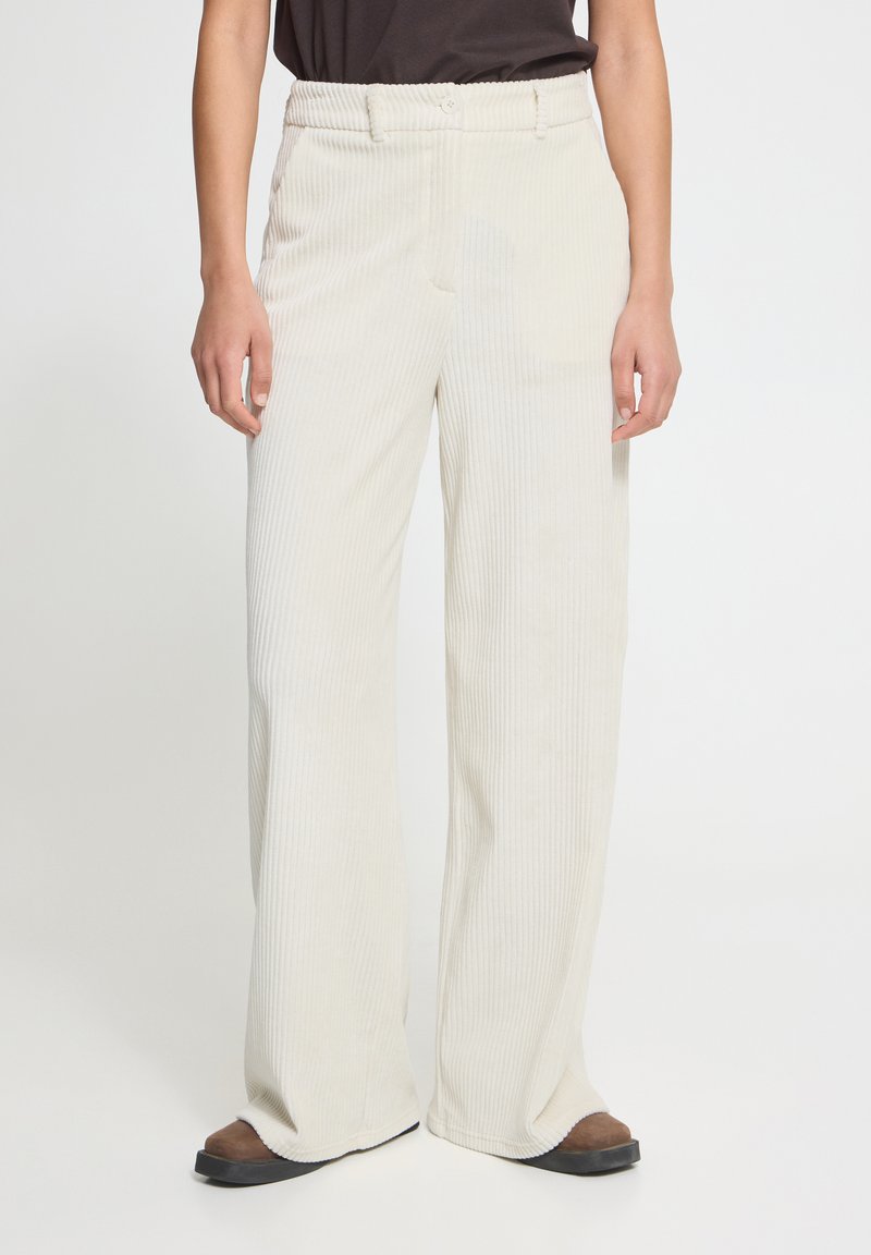 Personne portant un pantalon large en velours côtelé blanc cassé avec fermeture par bouton et zip, associé à des chaussures marron et un haut foncé.