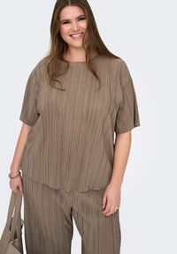 Χαμογελαστή γυναίκα με μακριά μαλλιά φοράει ταιριαστό φαρδύ τοπ και παντελόνι σε αποχρώσεις του ταουπέ, κρατώντας μπεζ τσάντα ώμου, σε ανοιχτό γκρι φόντο.