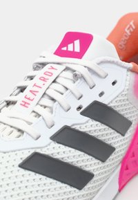 Scarpa atletica bianca con materiali in rete, dettagli grigi, marchio rosa sulla linguetta e suola traslucida a motivi. I lacci sono chiari.