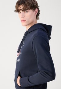 Sudadera de color azul marino hecha de una mezcla de algodón, que cuenta con un bolsillo tipo canguro, capucha con cordón y un diseño gráfico con acentos en rojo y blanco.