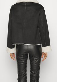 En person med en svart kort jacka med vita pälsmanchetter, i kombination med svarta läderbyxor, ses från bakom.