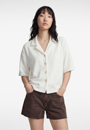Vrouw met zwart golvend haar die een wit overhemd met korte mouwen en donkere bruine denim shorts draagt, met haar handen in haar zakken.