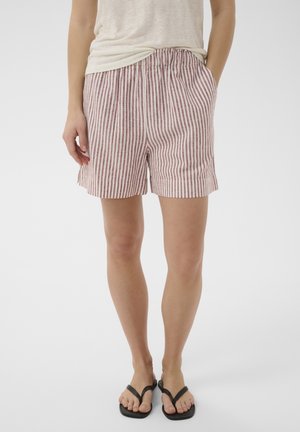 Person iført røde og hvide lodrette stribede shorts med lommer, en lysebeige top og sorte flip-flops, stående mod en hvid baggrund.