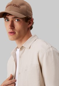 Camisa de botón color beige con textura sutil, que cuenta con un bolsillo en el pecho. Gorra marrón con el logotipo de "Calvin Klein" bordado en marrón oscuro.