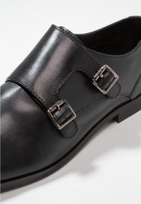 Scarpa da uomo in pelle nera con fibbie monaco argento su sfondo bianco, inclinata per mostrare i dettagli laterali e superiori.