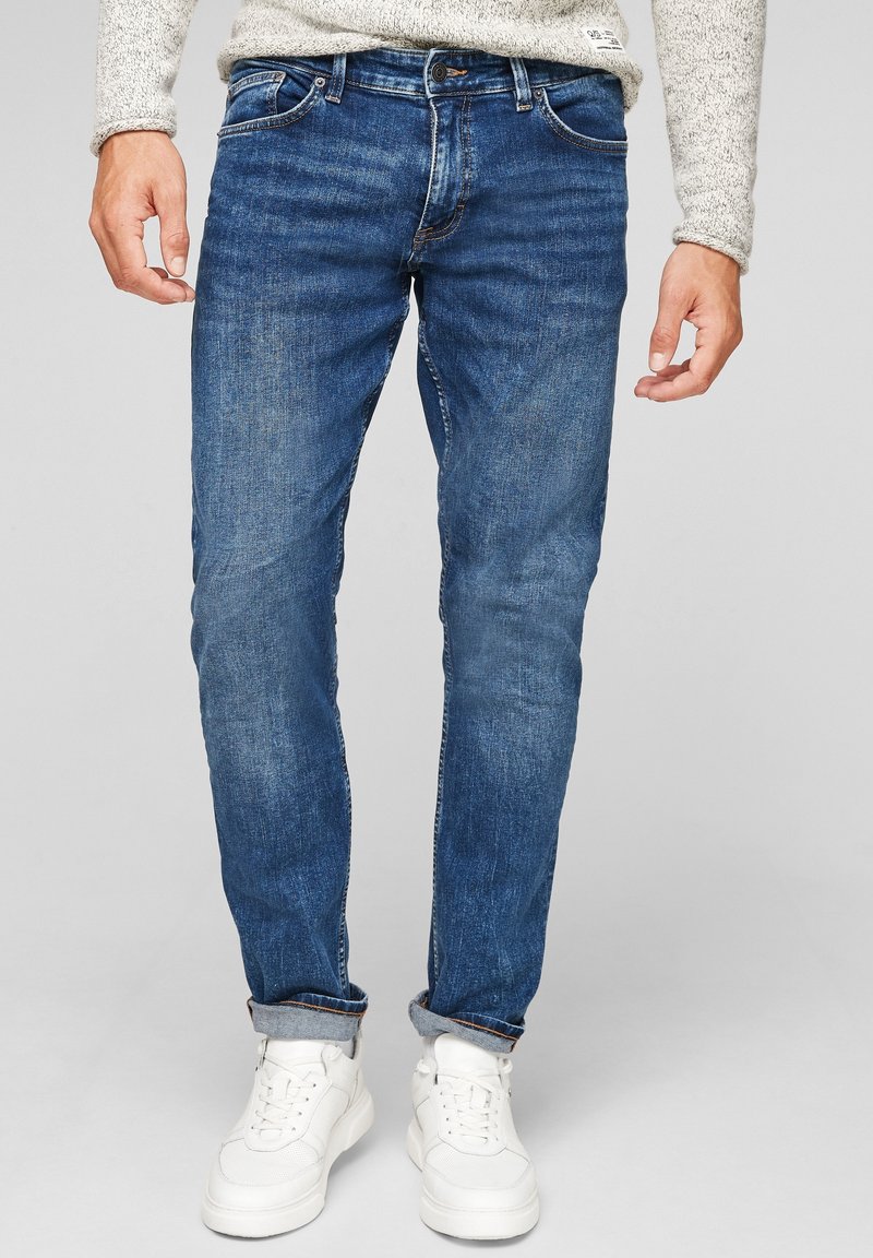 Jean en denim bleu porté avec des baskets blanches et un pull en maille gris clair, les ourlets du jean légèrement remontés.