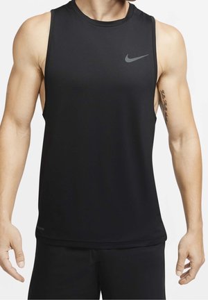 Muscularna osoba w czarnym sportowym, bezrękawnym topie Nike i czarnych krótkich spodenkach, pokazująca tatuaż na lewym górnym ramieniu.