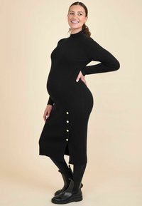 Abito premaman a coste nere con collo alto, spacco laterale e bottoni dorati. Abbinato a stivaletti neri, con uno sfondo beige.