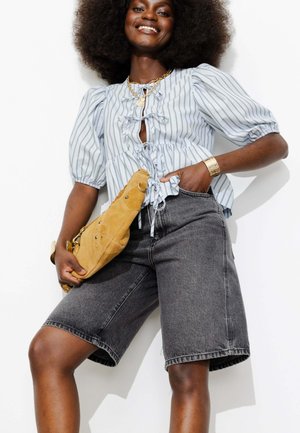 Femme souriante avec afro portant un chemisier à rayures et manches bouffantes, un short bermuda en jean gris, un bracelet en or, des colliers en couches, tenant un sac en daim beige.