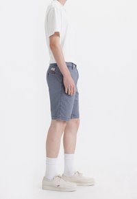 Shorts en denim gris avec une coupe régulière, poches latérales et un petit patch logo rouge à l'arrière. Associés à un t-shirt blanc à manches courtes et des chaussures claires.