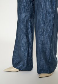 Wijde broekspijpen van een glanzende, gekreukte marineblauwe stof met een textuur, gecombineerd met beige pumps met spitse neus.