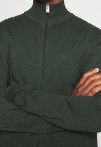 Grön ziptröja med hög krage, tillverkad av mjukt tyg. Har långa ärmar och en åtsittande design.