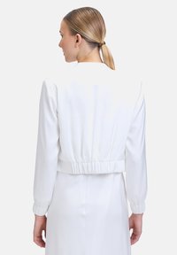 Veste blanche courte avec des manches longues, une taille élastique et une texture lisse. Vue de dos, sans motifs ni matériel visible.