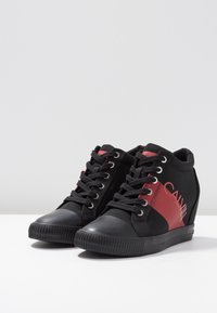 Svarta kilskor med svarta snören och röd diagonal rand som har delvis synlig texten "CALVIN" på sidan.